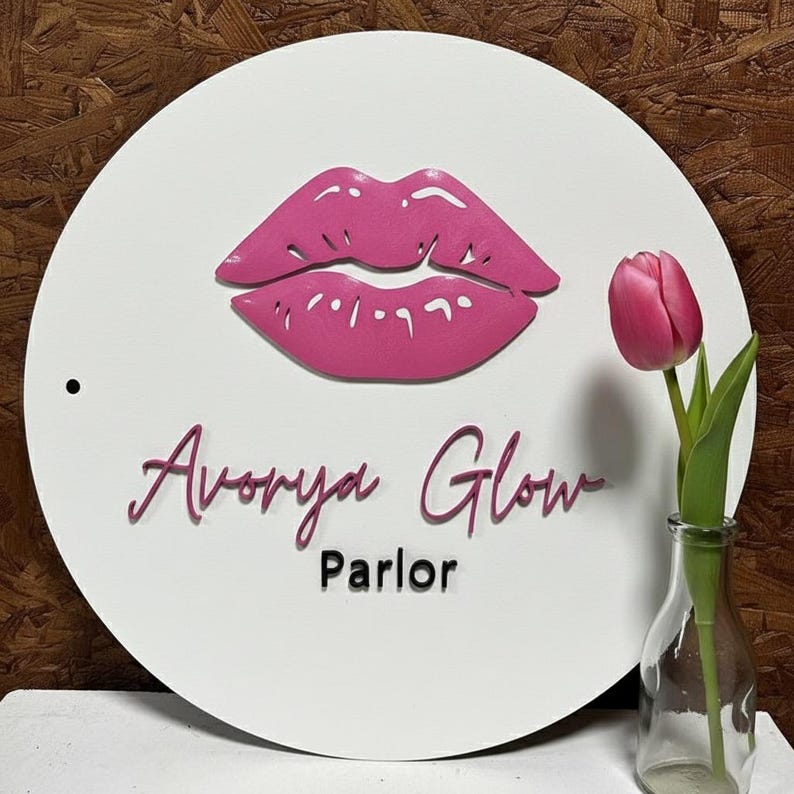 Pu&ograve; includere: Un cartello circolare bianco con una grafica in rilievo di labbra rosa nella parte superiore. Sotto le labbra, il testo recita Avorya Glow in un carattere corsivo rosa, con la parola Parlor in un carattere nero pi&ugrave; piccolo. Un foro &egrave; visibile.