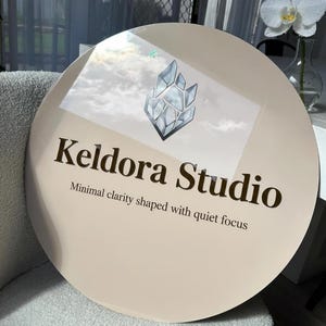 May include: A circular beige sign featuring a dark brown logo of a geometric crystal shape above the text Keldora Studio and the tagline Minimal clarity shaped with quiet focus in dark brown lettering on a smooth reflective surface.
