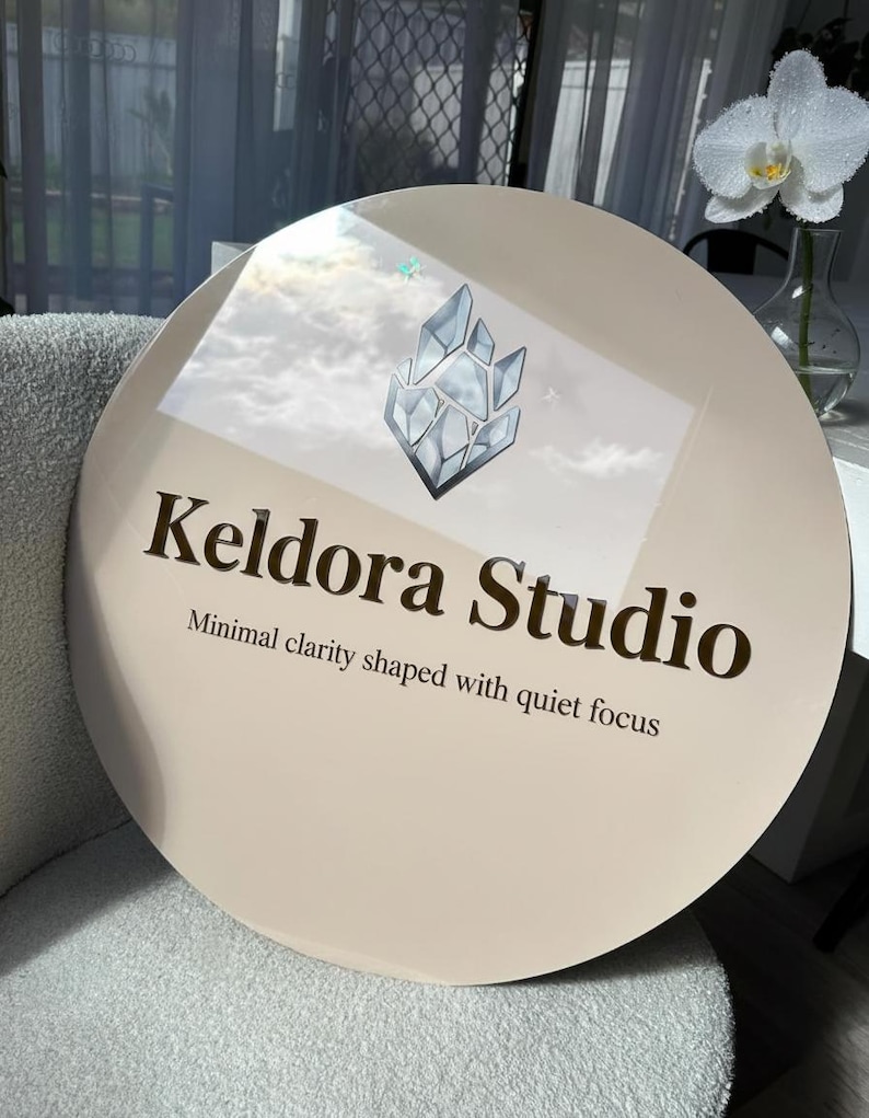 May include: A circular beige sign featuring a dark brown logo of a geometric crystal shape above the text Keldora Studio and the tagline Minimal clarity shaped with quiet focus in dark brown lettering on a smooth reflective surface.