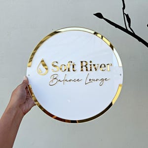 May include: A circular white sign with gold edging and lettering. The sign displays "Soft River Balance Lounge" in gold script. A gold teardrop shape is above the words. The sign is held by a hand.