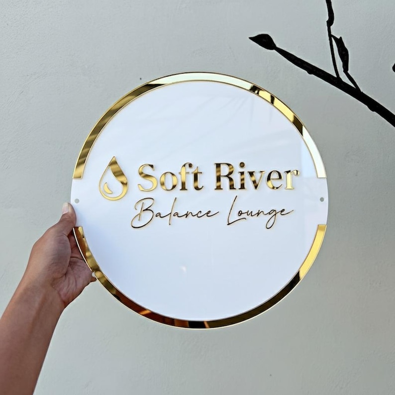 May include: A circular white sign with gold edging and lettering. The sign displays "Soft River Balance Lounge" in gold script. A gold teardrop shape is above the words. The sign is held by a hand.