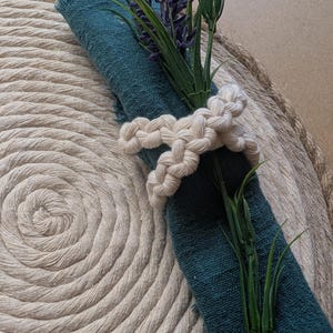 Puede incluir: Una servilleta de color turquesa está sujeta por un servilletero de macramé color crema, adornado con lavanda y vegetación artificial. La servilleta descansa sobre un salvamanteles de cuerda beige, creando una mesa rústica y elegante. La imagen muestra un primer plano de los elementos decorativos.