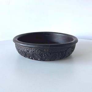 May include: A dark brown, round wooden bowl featuring carved relief animal figures around the outside. The bowl has a wide rim and a smooth interior. The carved animals include lions and other wildlife.