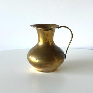 May include: A small, vintage brass jug with a rounded body and a curved handle. The jug has a spout for easy pouring and a warm, golden colour. The jug is likely used for serving liquids.