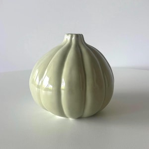 May include: A pale green ceramic vase, featuring a rounded form with vertical ribbed detailing. The vase has a small opening at the top and a glossy finish. This decorative item is suitable for displaying flowers or as a standalone accent.