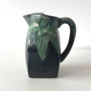 May include: A ceramic jug with a dark green and black glaze. The jug has a square base, a curved handle, and a spout. The top of the jug has a lid. The glaze has a textured, dripped appearance.