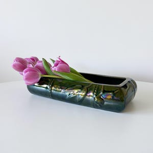 May include: A rectangular, dark green ceramic vase with iridescent highlights, holding several pink tulips. The vase has a textured, embossed design and sits on a white surface. The flowers add a pop of colour.