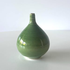 May include: A small, green ceramic vase with a rounded body and a narrow neck. The vase has a glossy finish and a subtle, textured pattern. The base is a lighter shade of green. This decorative vase is suitable for small floral arrangements.