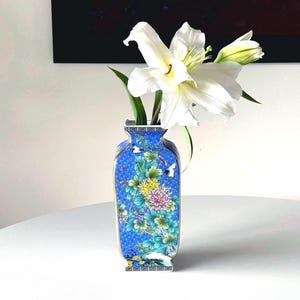 May include: A blue and white porcelain vase with floral designs and a white lily flower arrangement. The vase has a geometric pattern around the rim and base.