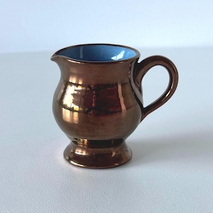 May include: A small, bronze-coloured creamer with a glossy finish. The interior is a contrasting light blue. It has a rounded body, a small spout, and a handle. The creamer sits on a small, circular base.