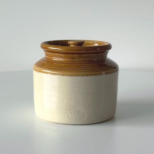 May include: A cream and brown stoneware jar with a lid. The jar has a cylindrical shape with a brown rim and lid. The lid features a small knob. This jar is likely for storing food or other items.