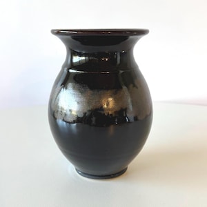 May include: A glossy black ceramic vase with a flared rim and a rounded body. The vase has a dark brown rim and a metallic silver band around the middle. The vase is a decorative item.