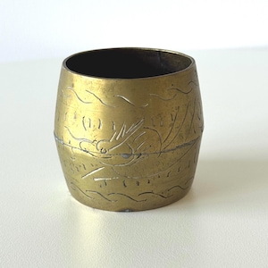 May include: A small, cylindrical brass container with etched dragon and wave designs. The container has a dark interior and a slightly barrel-shaped body. The brass has a warm, aged patina.