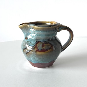 May include: A small, handcrafted ceramic jug with a rounded body and a handle. The jug features a glossy glaze in shades of teal, brown, and red. The design includes a spout for easy pouring. A decorative and functional piece for serving.