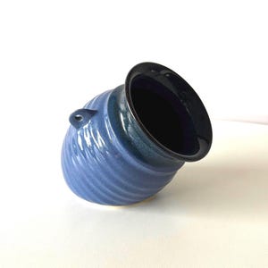 May include: A blue ceramic vase with a black rim and a small handle. The vase has a textured, ridged design and a dark interior. The vase is tilted, showcasing its shape and colour variations.