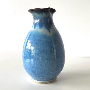 May include: A blue ceramic vase with a glossy finish. The vase has a rounded body and a flared, ruffled rim with a dark brown edge. The colour transitions from a lighter blue at the top to a deeper blue towards the base.