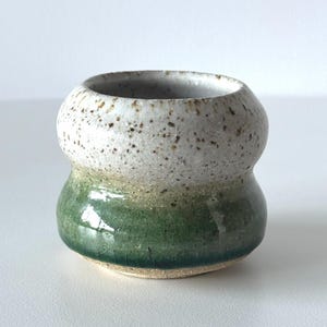May include: A small, handmade ceramic vessel with a distinctive form. The top section is speckled white, fading into a glossy green base. The interior is a light, neutral hue. The vessel is likely for decorative use.