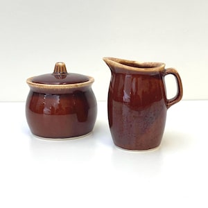 May include: A brown ceramic sugar bowl with a matching lid and a brown ceramic creamer jug. Both pieces feature a glossy, deep brown glaze with lighter tan-coloured edges along the rims and the handle of the jug.