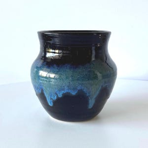 May include: A handcrafted ceramic vase with a glossy finish. The vase has a dark blue top that fades into a teal and blue glaze, creating a dripping effect. The vase has a rounded body and a slightly flared rim.