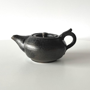 May include: A small, black ceramic teapot with a lid and handle. The teapot has a textured surface and a spout for pouring. The handle is a loop shape. The teapot is sitting on a white surface.