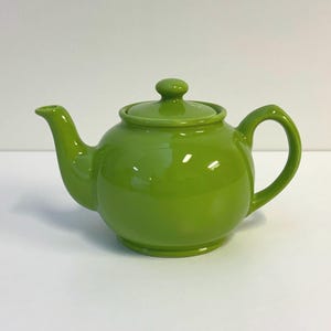 May include: A bright lime green ceramic teapot with a glossy finish. The teapot features a rounded body, a curved spout, a C-shaped handle, and a matching removable lid with a small knob handle on top. The design is simple and functional.