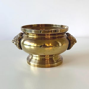 May include: A vintage brass bowl with a wide, open top and a rounded body. The bowl features decorative lion head handles on either side. The base is a pedestal design with horizontal ridges. The brass has a polished, reflective finish.