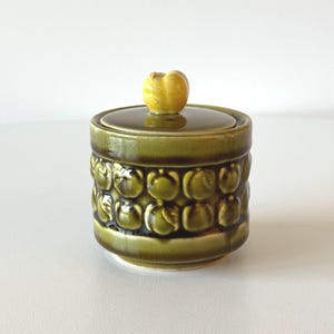 May include: A vintage green ceramic jar with a lid and a yellow knob. The jar features a repeating pattern of embossed shapes. The lid is a darker shade of green. A decorative kitchen storage container.