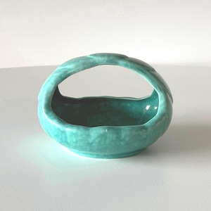 May include: A small, turquoise ceramic basket with a curved handle. The basket has a rounded base and a textured handle, and is likely used for decorative purposes.