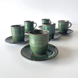 May include: A set of six green ceramic espresso cups and saucers. The cups have a dark green exterior with a lighter green interior and a small handle. The saucers are dark green with a slightly raised edge. The set is on a white surface.