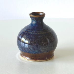 May include: A small, handcrafted ceramic vase with a rounded body and a narrow neck. The vase features a glossy, dark blue glaze with brown accents at the base and rim. The vase is approximately 10 cm tall.