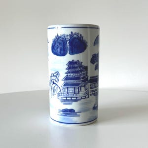 May include: A white ceramic cylindrical vase with a blue and white design of a pagoda and landscape. The vase is decorated with a traditional Chinese style pattern.