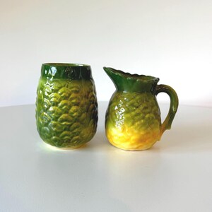 May include: A set of two green and yellow ceramic pineapple-shaped containers. The larger container is a jug with a handle, and the smaller container is a cup.