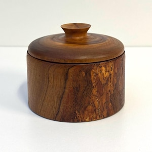 May include: A cylindrical wooden container with a matching lid featuring a small, centred knob. The exterior displays a natural brown wood grain pattern with a rough, textured bark-like section on one side, showcasing organic variations in colour.