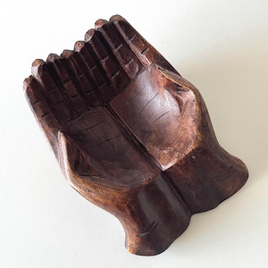May include: A carved wooden bowl shaped like cupped hands. The bowl is made from dark brown wood, showing the grain and texture. The fingers are detailed, and the bowl has a smooth, polished finish. Suitable for holding small items.