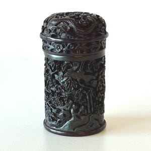 May include: A dark brown, cylindrical container with detailed relief carvings. The lid and body feature intricate patterns of plants, animals, and possibly mythical figures. The container has a rounded base and a matching lid, suggesting it is a decorative storage item.
