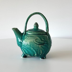 May include: A small, teal green ceramic teapot with a lid and a handle. The teapot has a textured surface and three small feet.