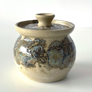 May include: A handmade ceramic jar with a lid. The jar is round with a beige base and a lid with a small handle. The jar's surface features a mottled pattern of blue, brown, and gold hues, creating a unique, artistic appearance. The jar is suitable for storing small items.