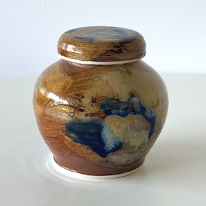 May include: A ceramic jar with a lid. The jar is a rounded shape with a brown base and a lighter brown body. Blue and tan accents are visible on the body. The lid is brown with blue accents. The jar sits on a white surface.