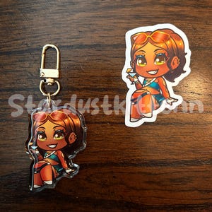 May include: A clear acrylic keychain and sticker featuring a cartoon character with tan skin, brown hair, and sunglasses. The character is wearing a blue top and orange bottoms, holding a cocktail. The keychain has a gold-colored clasp. The text "Stardustkinn" is visible.