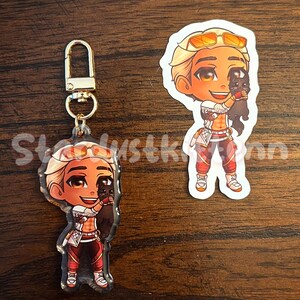 May include: Two items: a keychain and a sticker. Both feature a cartoon character with blonde hair, orange sunglasses, and red and white clothing, holding a black cat. The keychain has a gold clasp. The sticker is a cut-out of the character.