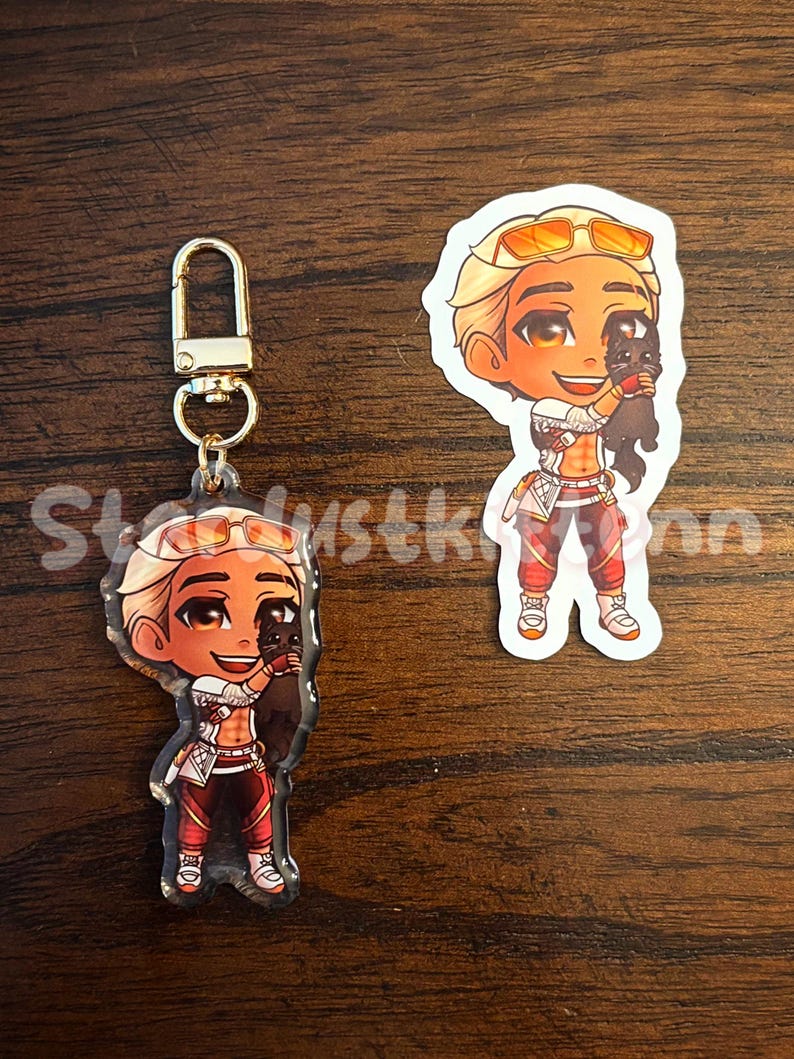 May include: Two items: a keychain and a sticker. Both feature a cartoon character with blonde hair, orange sunglasses, and red and white clothing, holding a black cat. The keychain has a gold clasp. The sticker is a cut-out of the character.