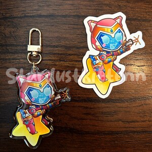 Op de afbeelding: Een sleutelhanger en sticker met een cartoonfiguur met katachtige kenmerken, gekleed in een futuristisch kostuum en met een wapen. Het personage staat tegen een gele ster. De sleutelhanger heeft een goudkleurige sluiting. De sticker heeft een vergelijkbaar ontwerp.