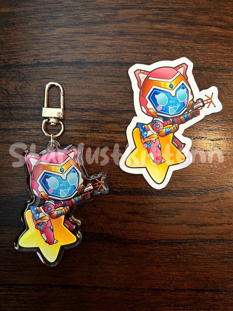 Op de afbeelding: Een sleutelhanger en sticker met een cartoonfiguur met katachtige kenmerken, gekleed in een futuristisch kostuum en met een wapen. Het personage staat tegen een gele ster. De sleutelhanger heeft een goudkleurige sluiting. De sticker heeft een vergelijkbaar ontwerp.
