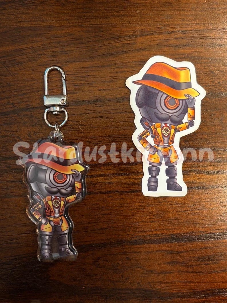 May include: Two robot-themed accessories: a keychain and a sticker. The keychain features a robot character with a hat and a mustache. The sticker shows the same robot character. Both have orange and yellow accents.