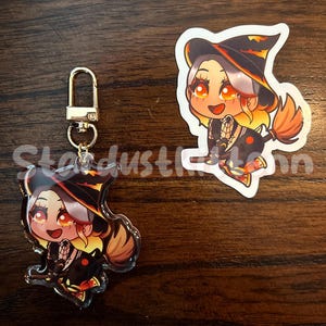 May include: A keychain and sticker featuring a cartoon witch with orange and yellow hair, wearing a black hat and dress. The keychain has a gold clasp. The sticker is white with a black border. The text "Stardust" is partially visible.