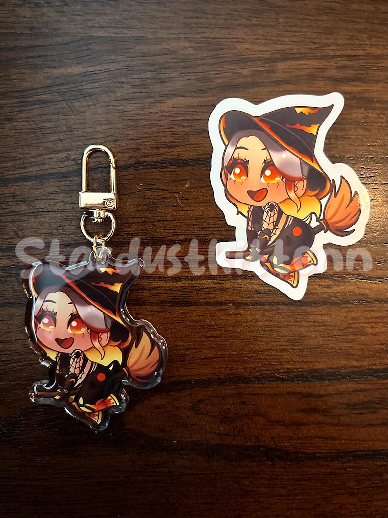 May include: A keychain and sticker featuring a cartoon witch with orange and yellow hair, wearing a black hat and dress. The keychain has a gold clasp. The sticker is white with a black border. The text "Stardust" is partially visible.