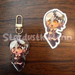 May include: An acrylic charm and sticker featuring a cartoon character with blonde hair, gold eyes, and a black outfit. The charm has a gold clasp. The sticker is a die-cut shape. The text "StardustKinn" is visible.