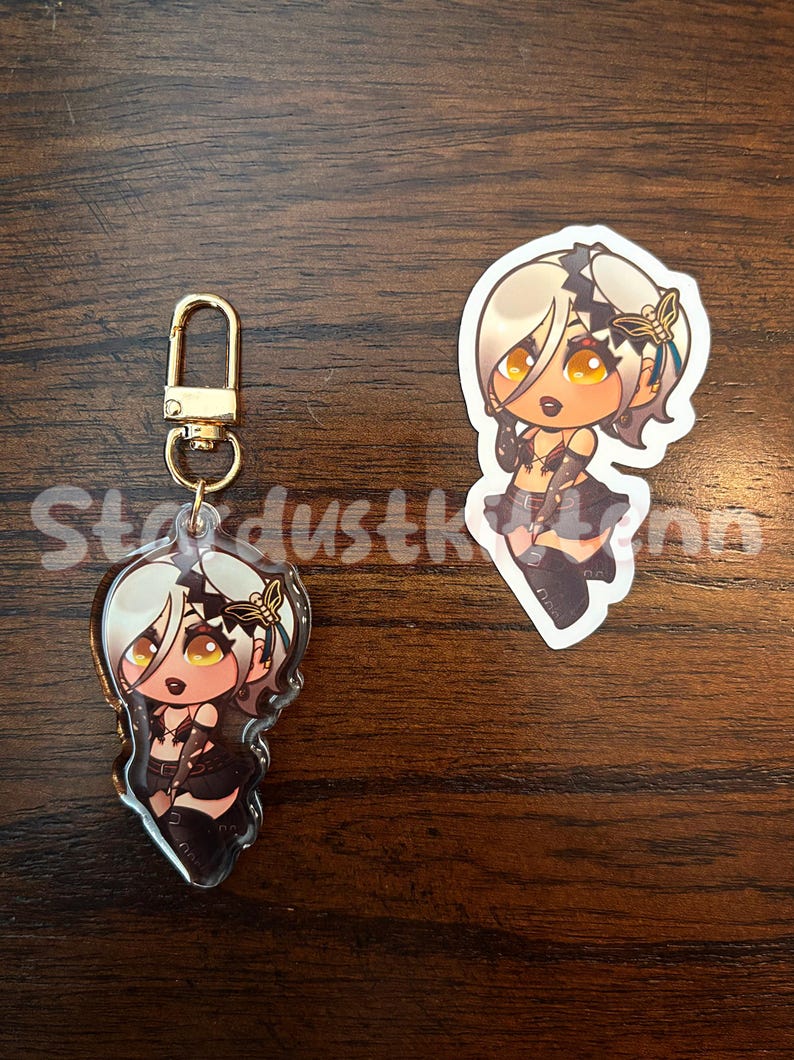 May include: An acrylic charm and sticker featuring a cartoon character with blonde hair, gold eyes, and a black outfit. The charm has a gold clasp. The sticker is a die-cut shape. The text "StardustKinn" is visible.