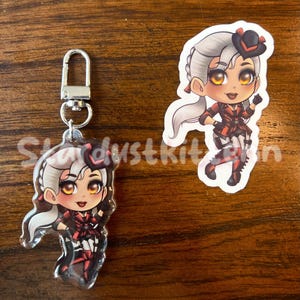 K&ouml;nnte beinhalten: Zwei Charms mit Anime-Charakteren. Einer ist ein Schl&uuml;sselanh&auml;nger mit einem silbernen Verschluss, der andere ein Aufkleber. Beide zeigen einen Charakter mit grauem Haar, einem schwarzen Hut und einem rot-schwarzen Outfit. Der Text "Stardustkit" ist teilweise sichtbar.