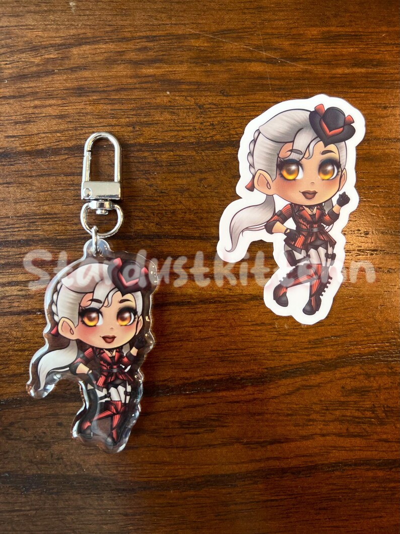 K&ouml;nnte beinhalten: Zwei Charms mit Anime-Charakteren. Einer ist ein Schl&uuml;sselanh&auml;nger mit einem silbernen Verschluss, der andere ein Aufkleber. Beide zeigen einen Charakter mit grauem Haar, einem schwarzen Hut und einem rot-schwarzen Outfit. Der Text "Stardustkit" ist teilweise sichtbar.
